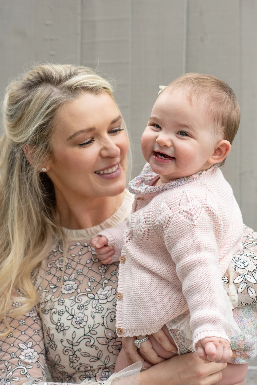 Baby girl in pink cardigan laughing as she is held by her mother smiling on.