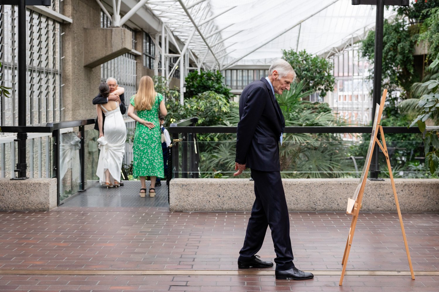 An older gentleman studies the seating plan whilst in the distance the bride hugs another guest.