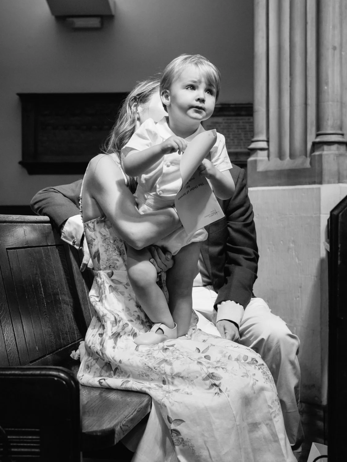 Baby brother standing on his mother's lap and looking up to his brother in the pulpit doing a reading
