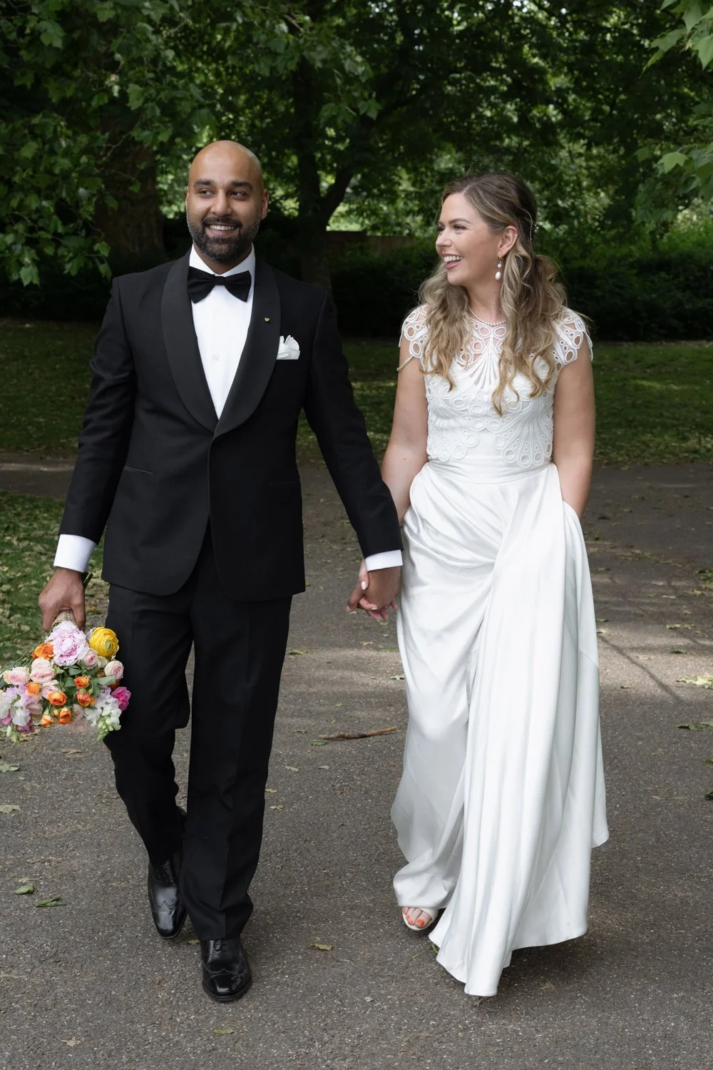 Bride and Groom walking along leafy path with groom carrying the bouquet