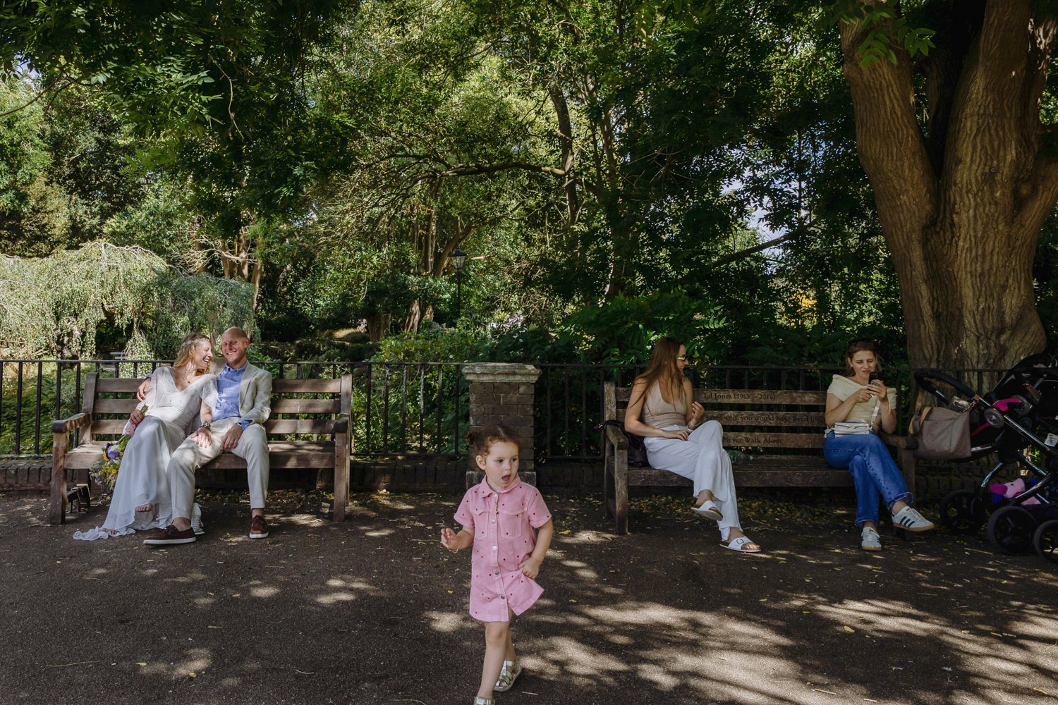 bride and groom on bench to left, and a child in a pink dress running into shot with two mothers on the bench the other side.