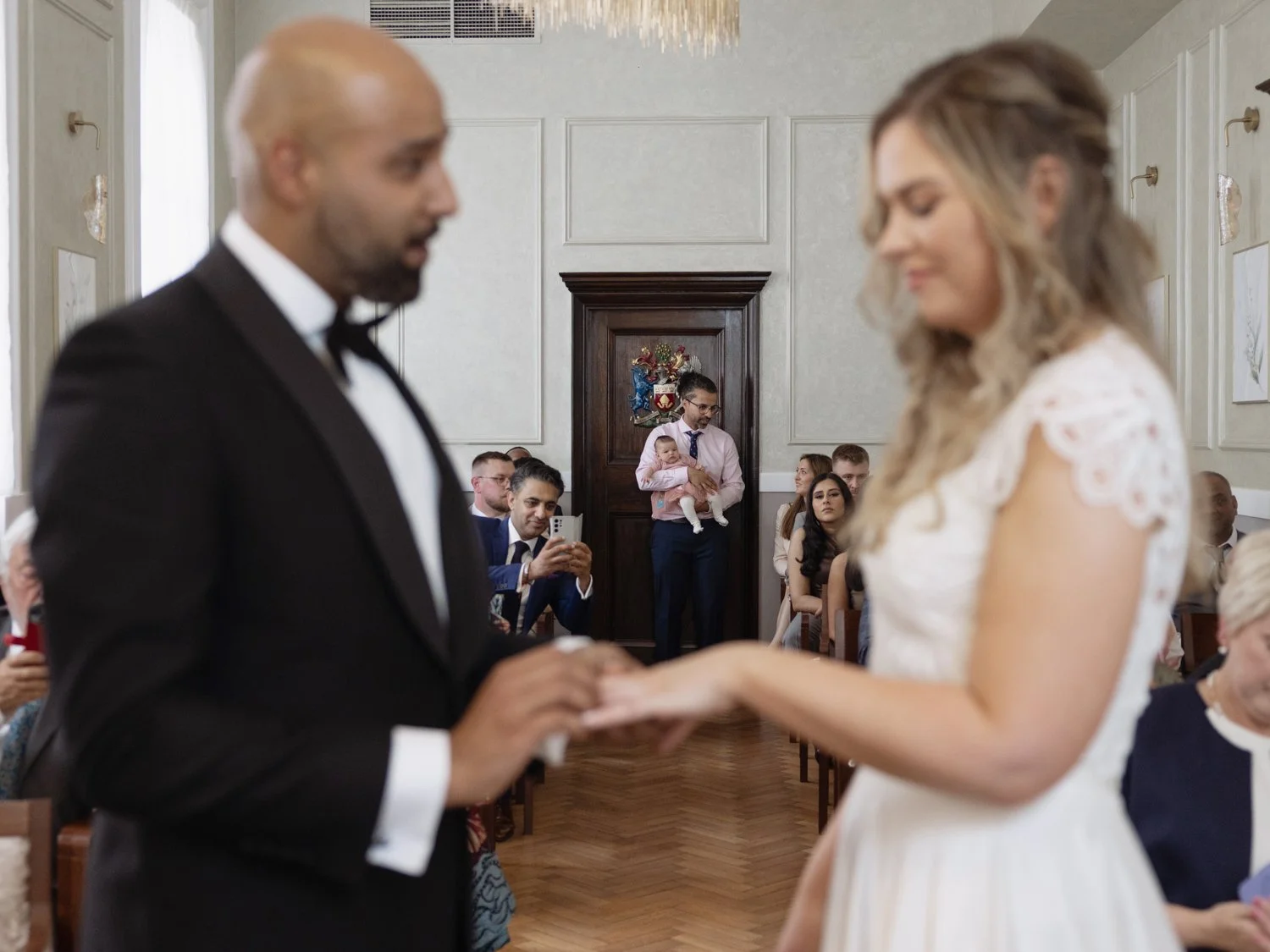 Bride and Groom exchanging rings in the foreground with a guest holding a baby at the back of the room in focus