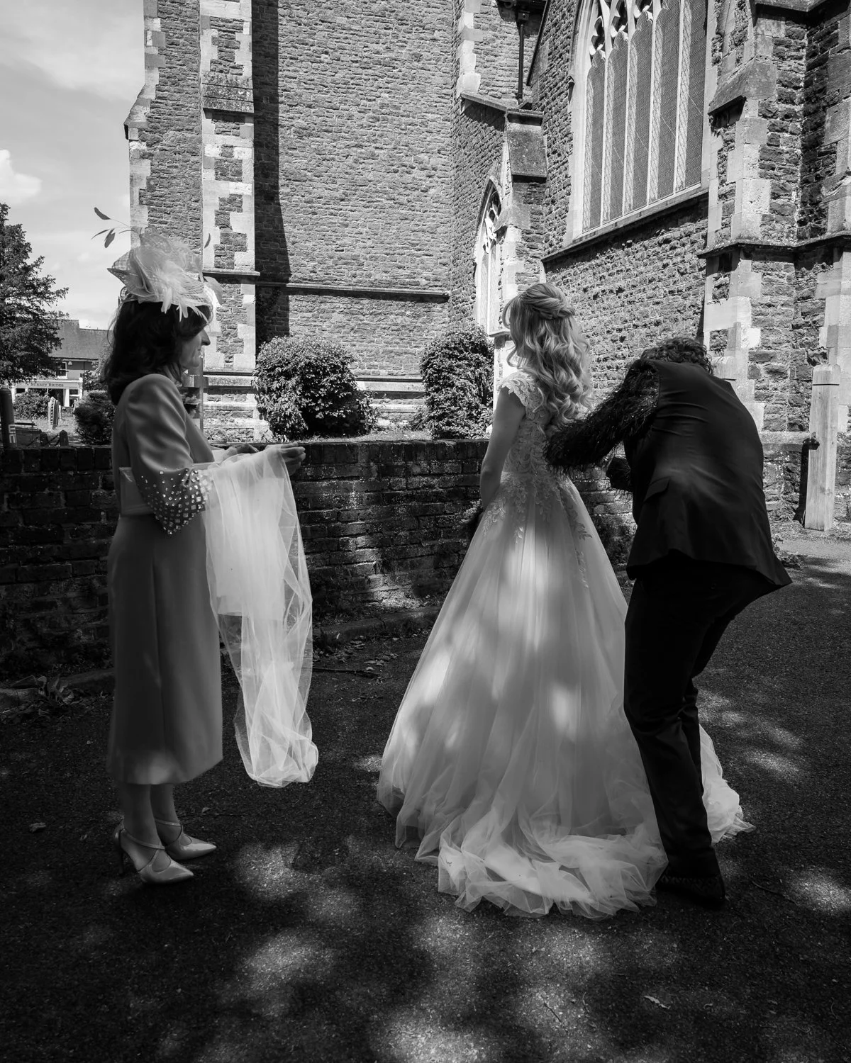Bride having her dress adjusted outside the church with her mother standing by ready with the veil.