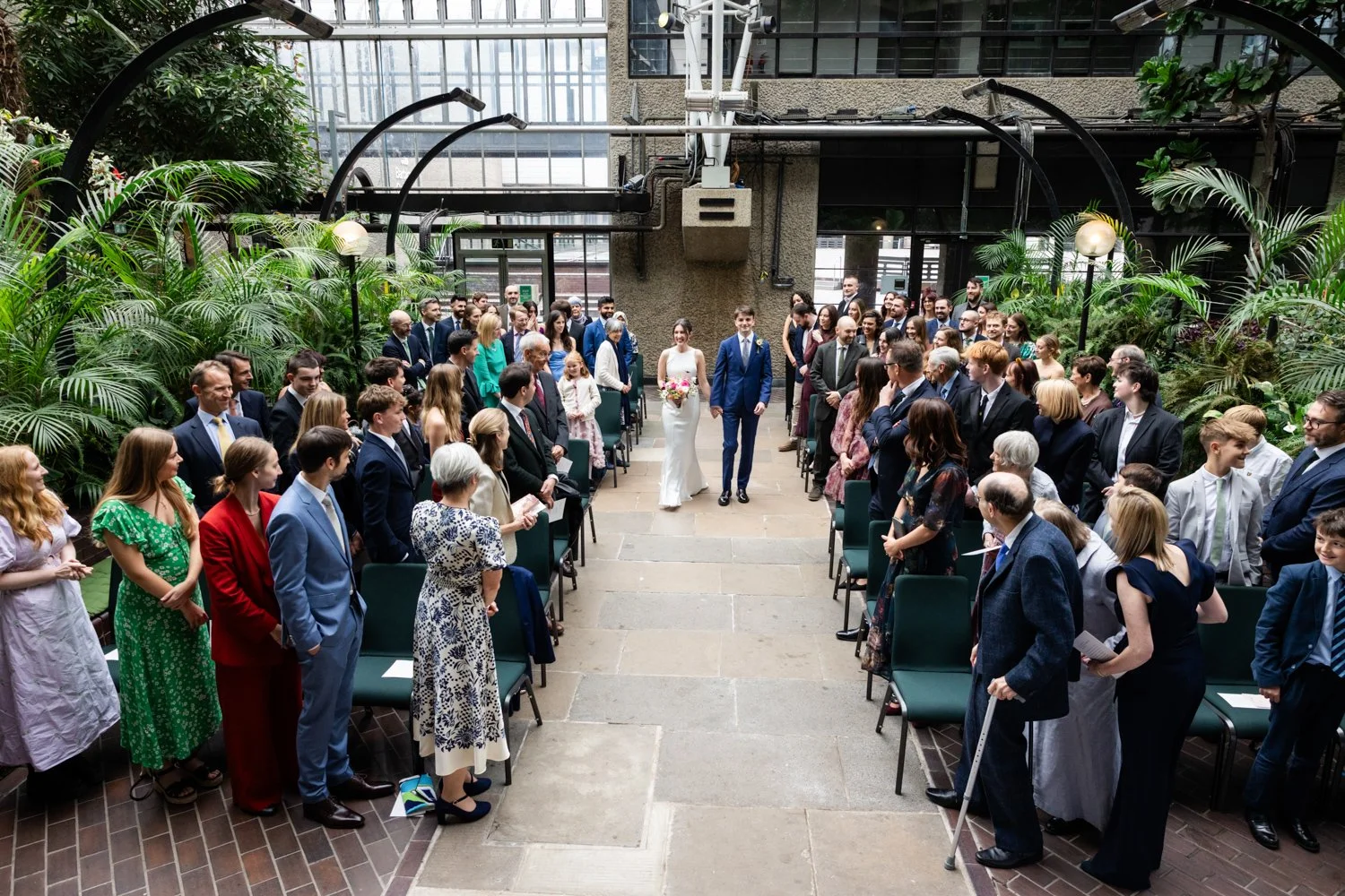 Bride and Groom travel up the aisle together in the Barbican Conservatory with guests turning around and looking on.