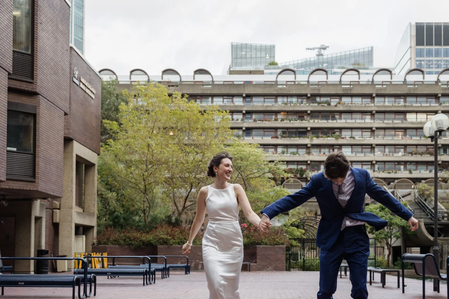 Bride and Groom holding hands running through the Barbican with the wind blowing and the Groom looking down and the Bride laughing.