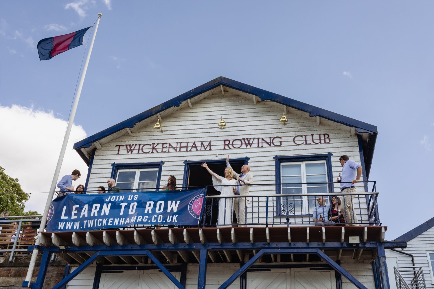 Bride and Groom waving to rowers on the river from the balcony of Twickenham Rowing Club.