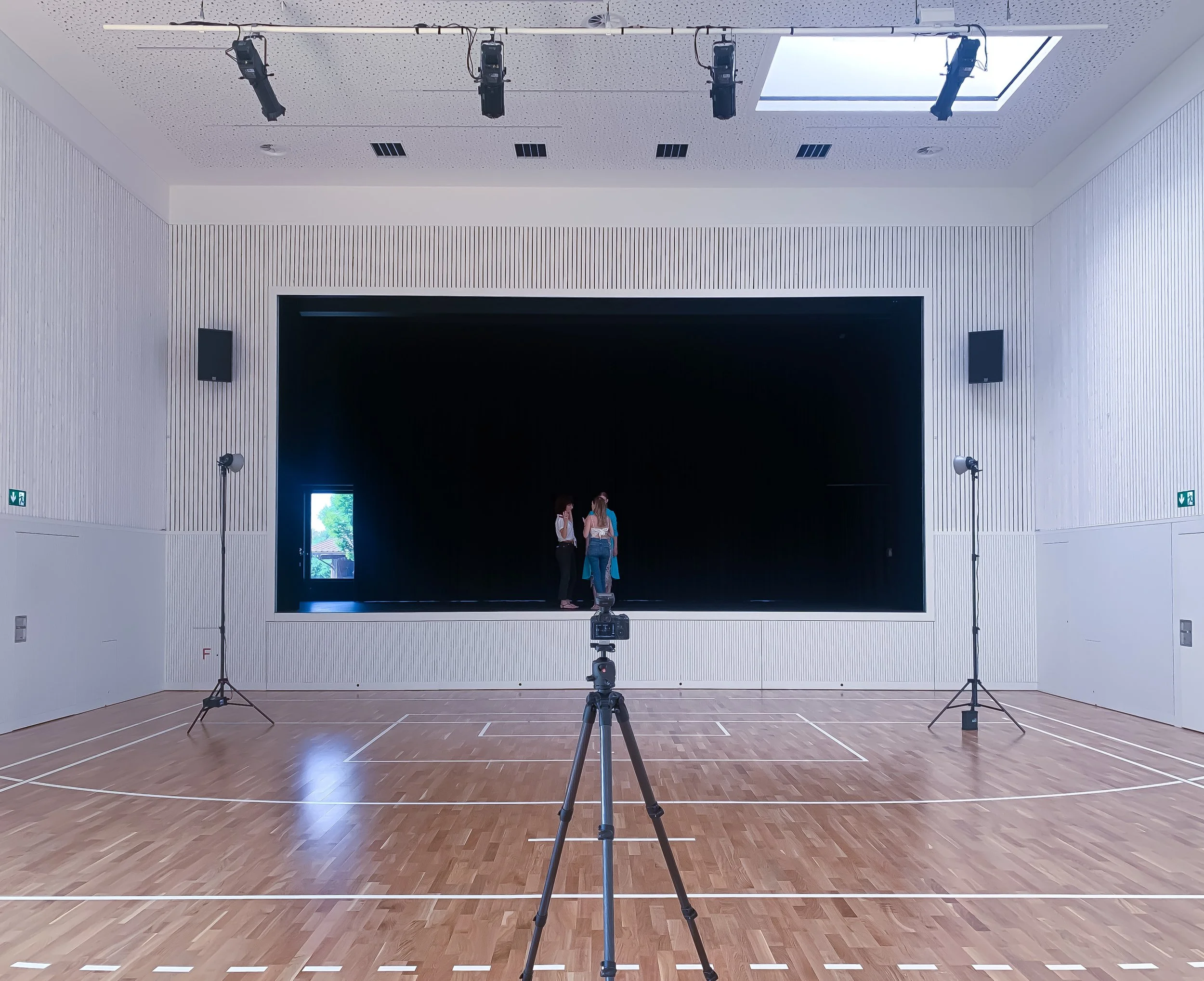 Zwei Personen stehen auf einer Bühne in einem großen, leeren Raum, umgeben von Studioausrüstung und Scheinwerfern.