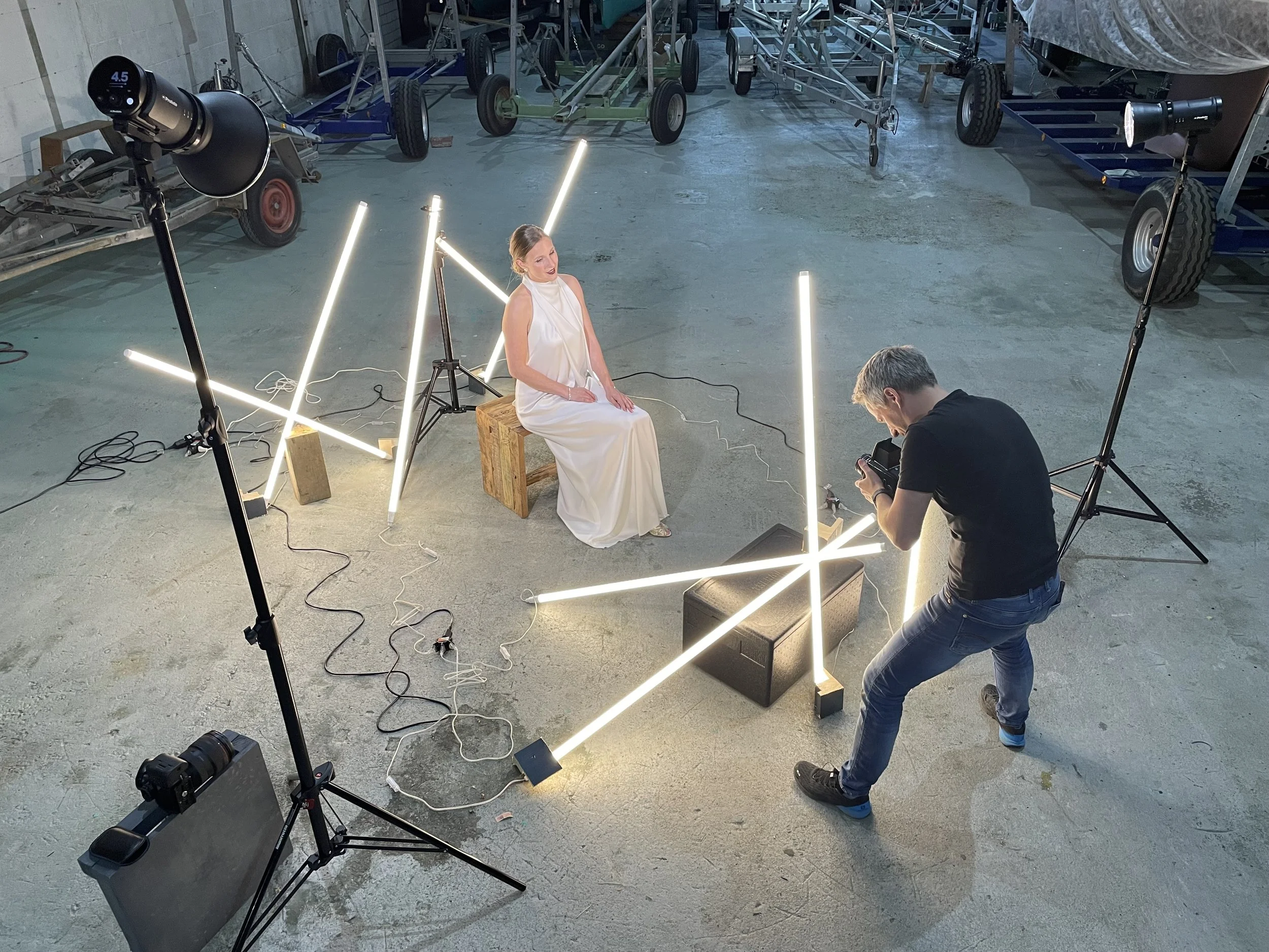 Ein Foto von einem Fotoshooting in einem Industriegebäude, bei dem eine Frau in einem weißen Kleid sitzt. Es gibt mehrere Lichtquellen, einen Fotografen, der ein Foto macht, und zwei große Scheinwerfer. Es sind auchMobile Arbeitsgeräte und Werkzeuge 