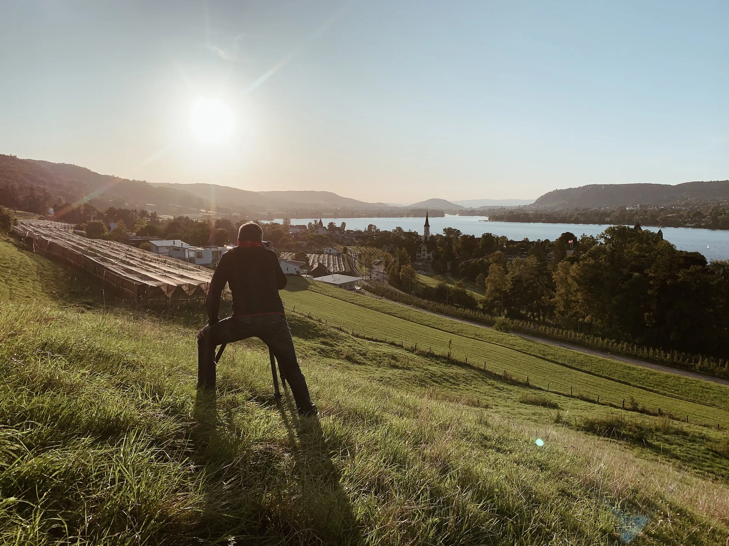 Person mit Kamera fotografiert die Landschaft bei Sonnenuntergang, mit Fluss, Hügel und Kirche im Hintergrund.