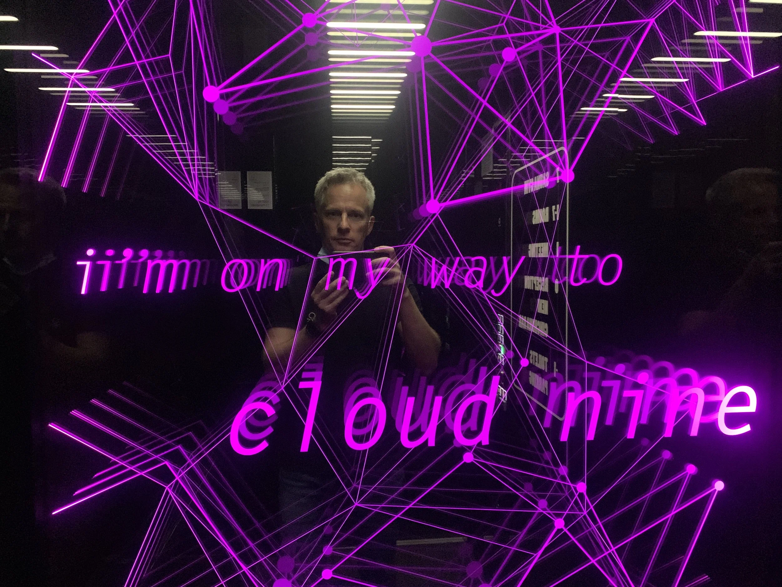 Reflexion eines Mannes in einer Neon-Leuchtwerbung mit dem Text 'iimm on my way to cloud mine' und pinken Netzlinien