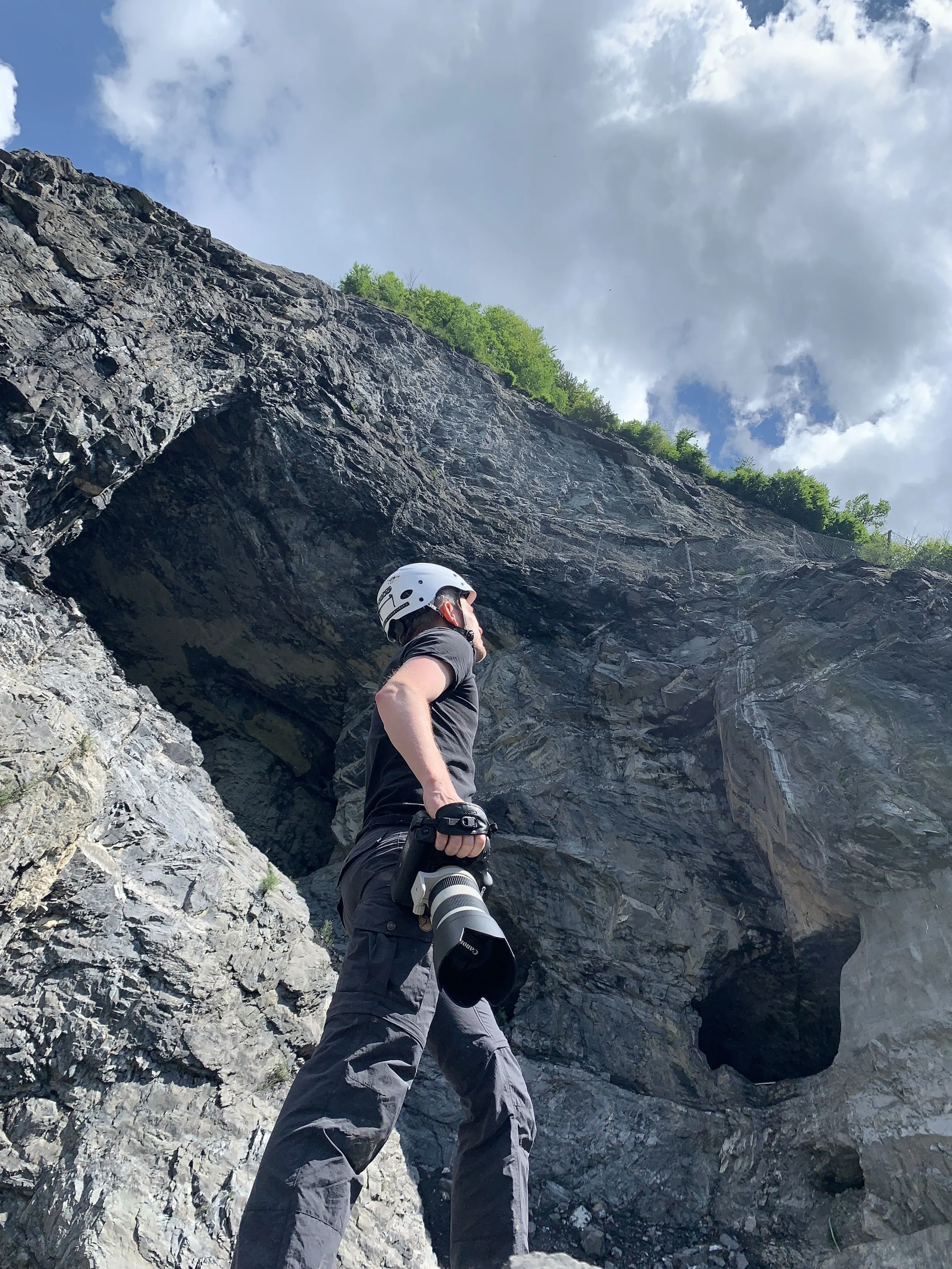 Ein Fotograf mit Helm steht vor einer Felswand mit einer Höhle oder einem Tunnel im Hintergrund, unter blauem Himmel mit Wolken.