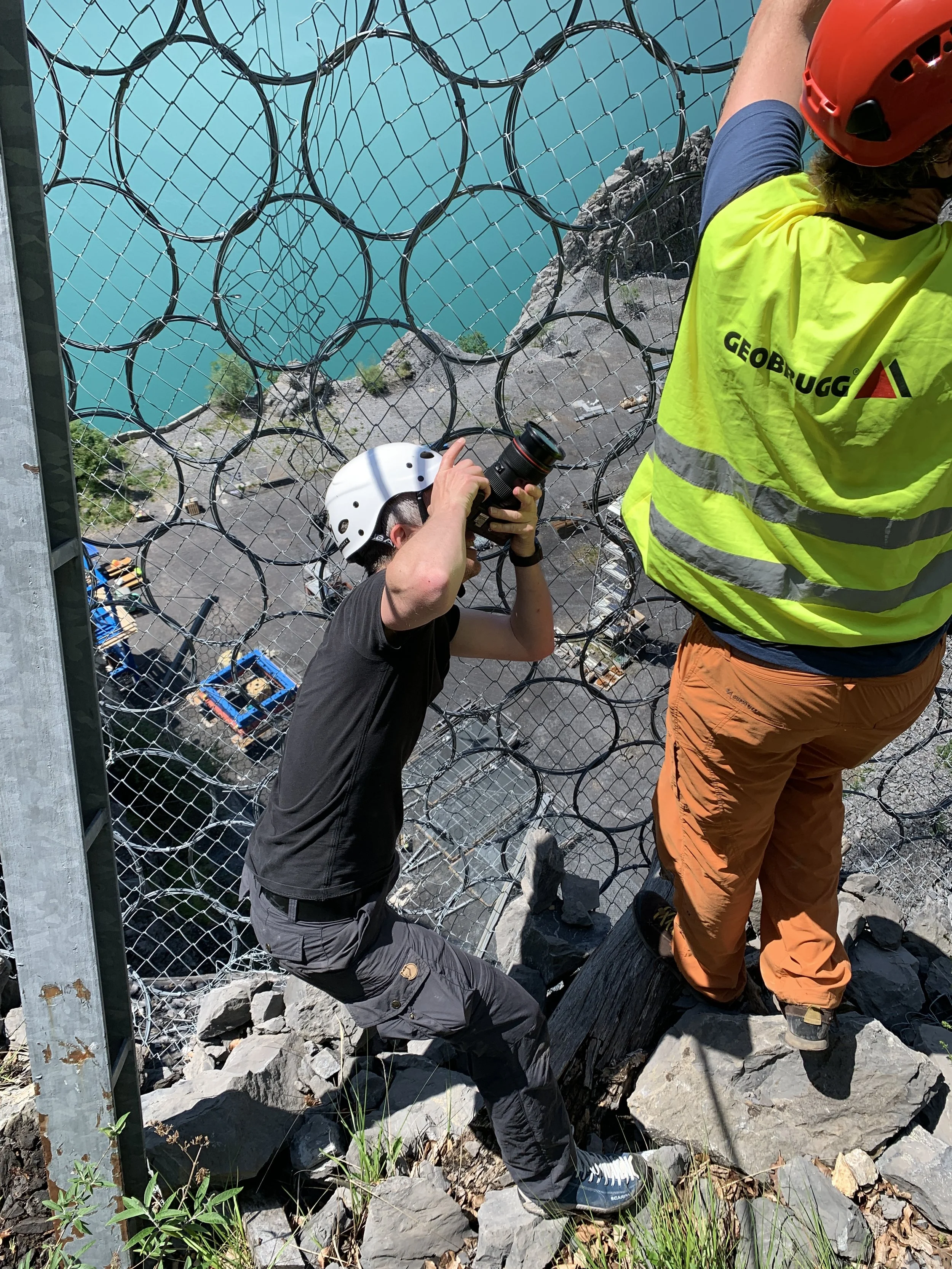 Zwei Menschen auf einem Felsen neben einem Sicherheitszaun, einer fotografiert, während der andere hinter dem Zaun steht, mit Blick auf einen Fluss oder See.