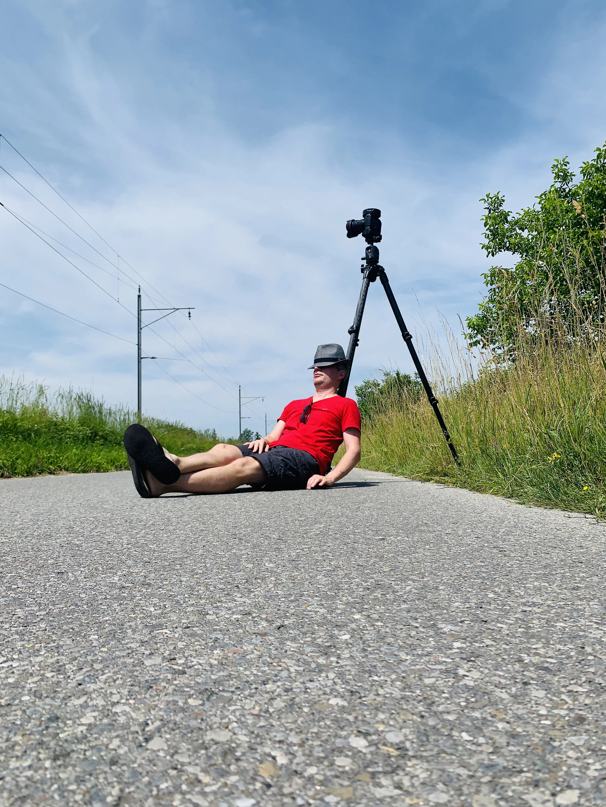 Mann in rotem T-Shirt und schwarzen Shorts liegt auf einer Straße, trägt einen Hut und hat ein Kamerastativ mit Kamera aufgestellt, Sonnenbrille hängt am T-Shirt, Himmel ist blau mit Wolken, umgeben von Gräsern und Büschen.