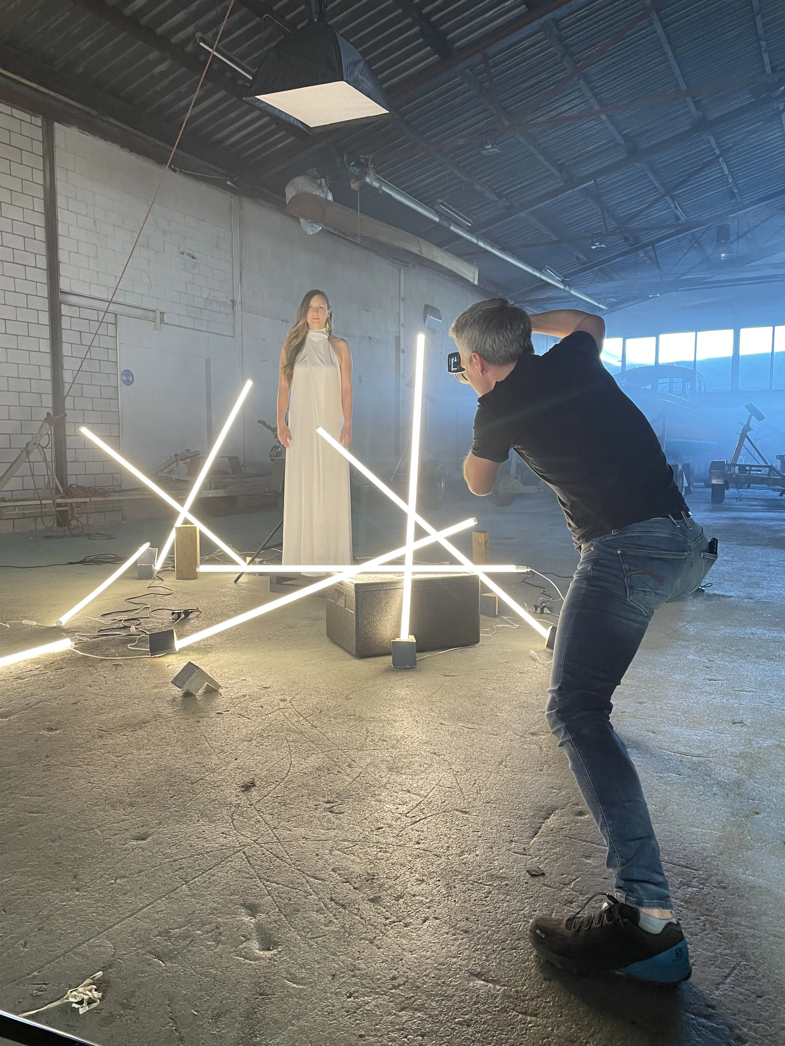Fotograf macht ein Fotoshooting in einer Fabrikhalle mit Beleuchtung aus Neonröhren, während die Modenschönheit in einem langen weißen Kleid posiert.