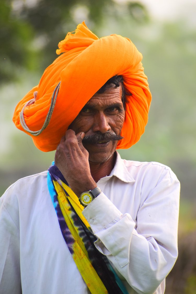 A man in a bright orange turban and white shirt, holding his hand to his face, outdoors.