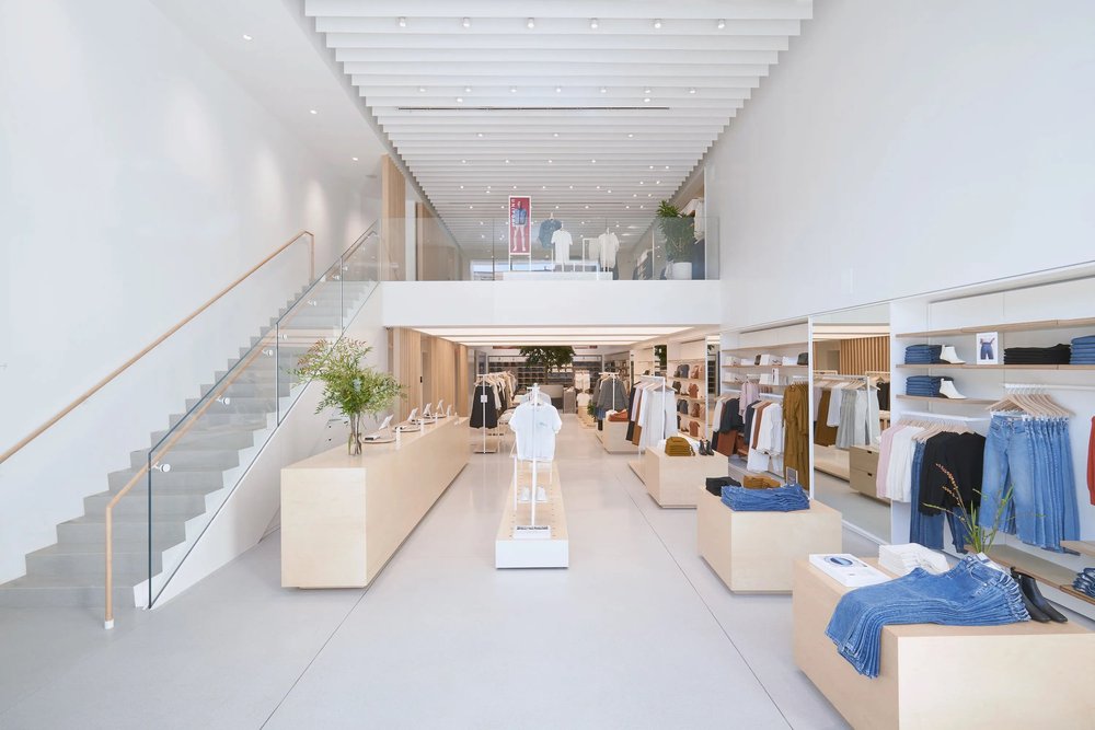 A bright, minimalist clothing store with racks of clothes, shelves of folded garments and a staircase to an upper level.