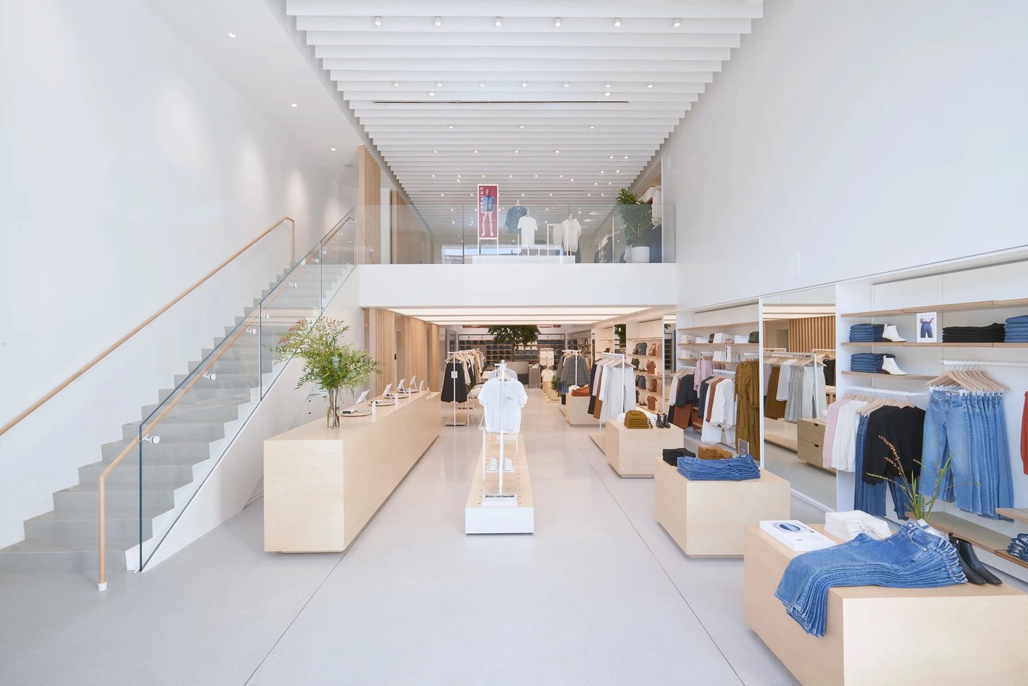 A bright, minimalist clothing store with racks of clothes, shelves of folded garments and a staircase to an upper level.
