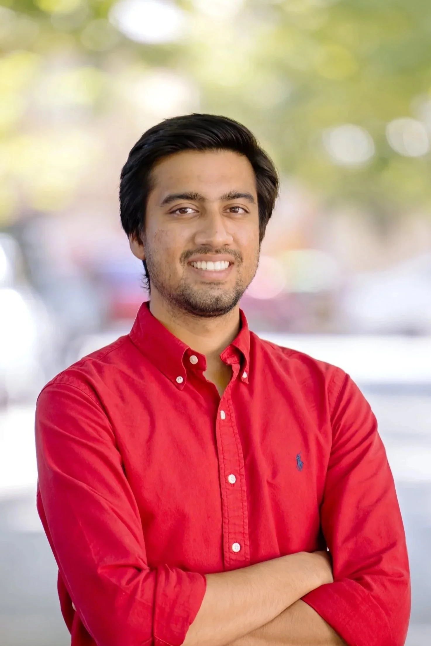 A young man with dark hair and light skin, wearing a red button-up shirt with sleeves rolled up, smiling with arms crossed, outdoors with blurred trees and cars in the background.