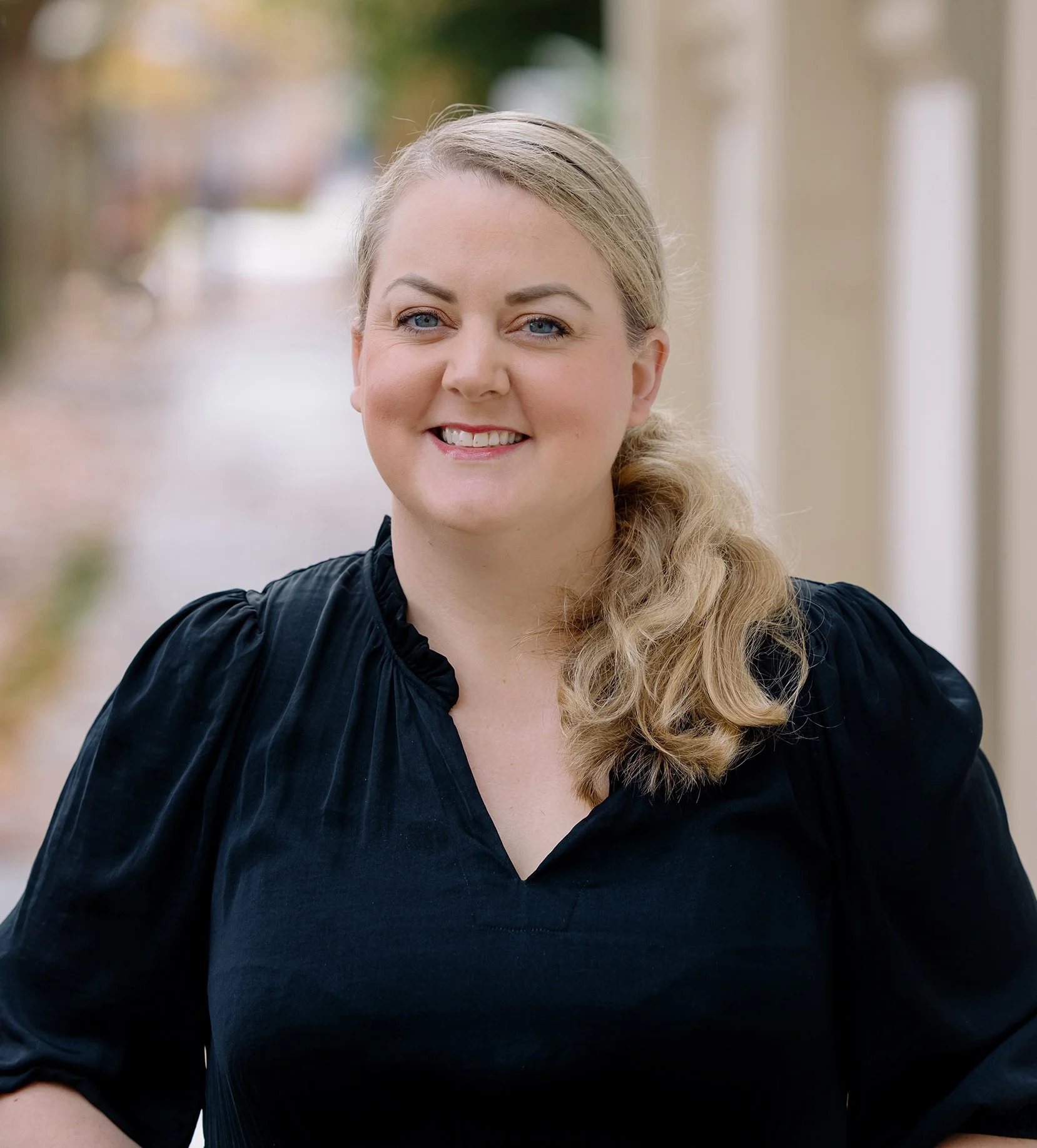 A woman with blonde hair in a ponytail, wearing a black blouse, smiling outdoors with a blurred background of trees and buildings.