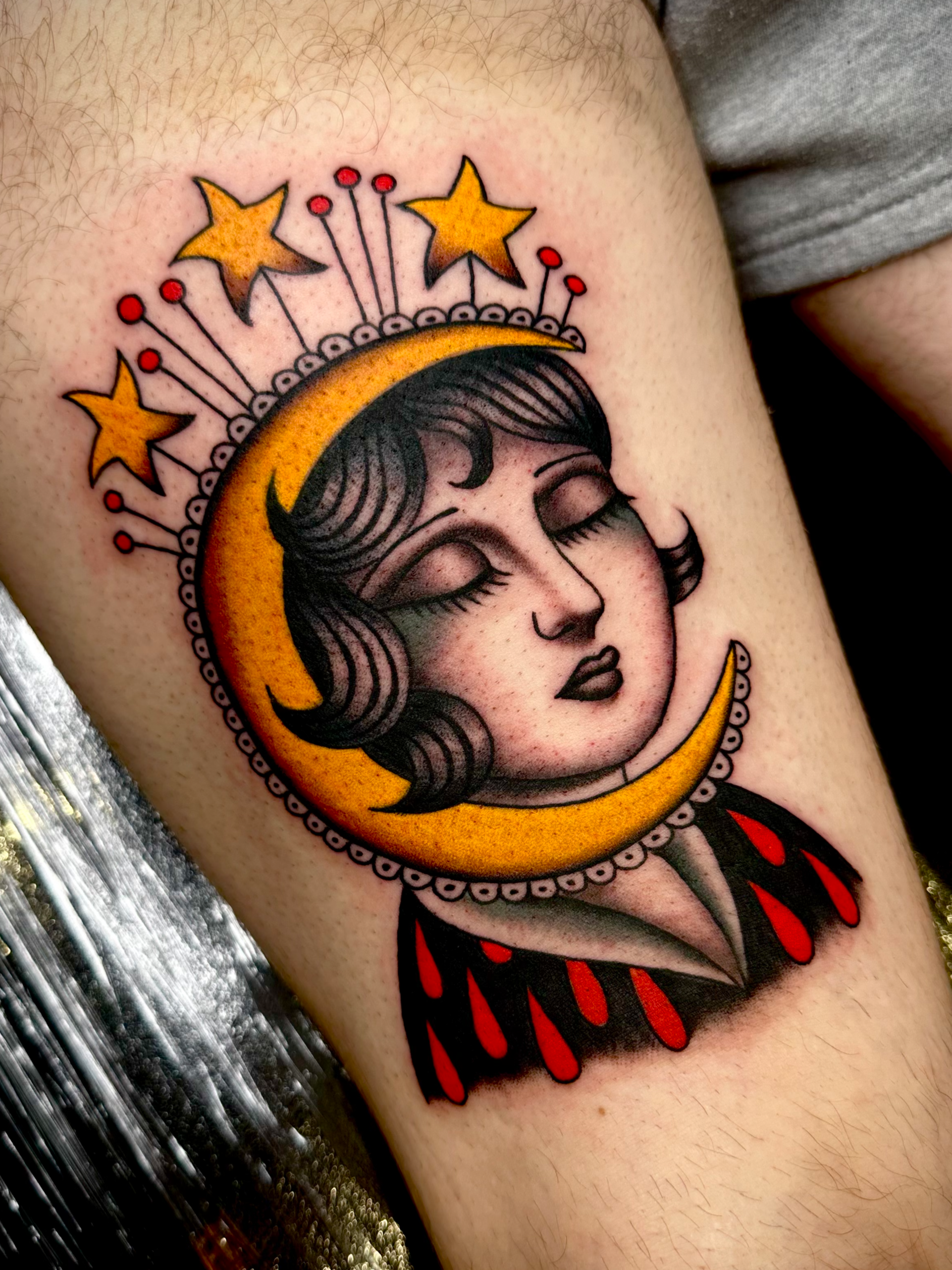 Tattoo of a woman's face with closed eyes, dark hair, and dark lipstick, surrounded by a crescent moon with stars and red accents.