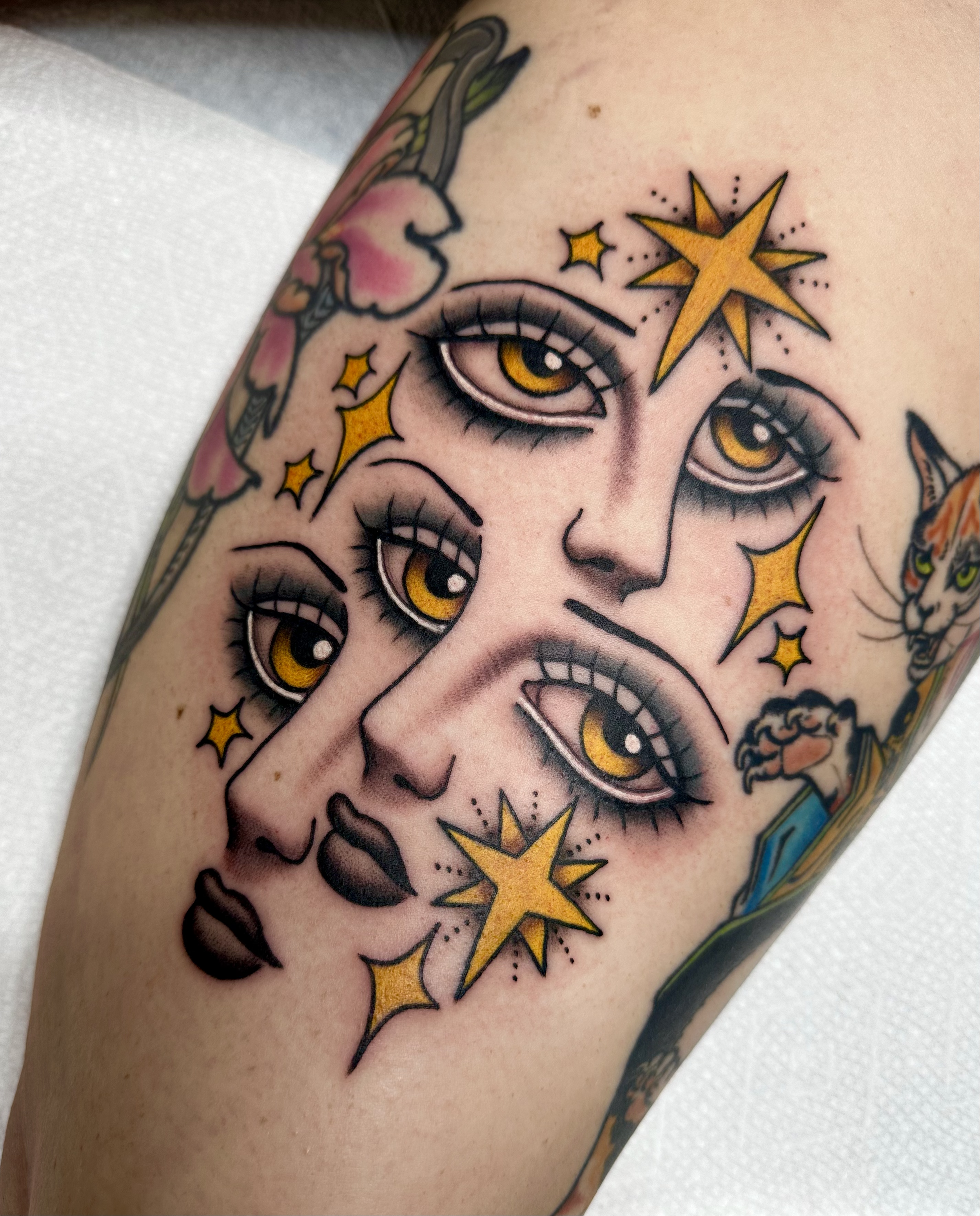 Tattoo of a woman's face with multiple eyes, surrounded by yellow stars and a colorful background.