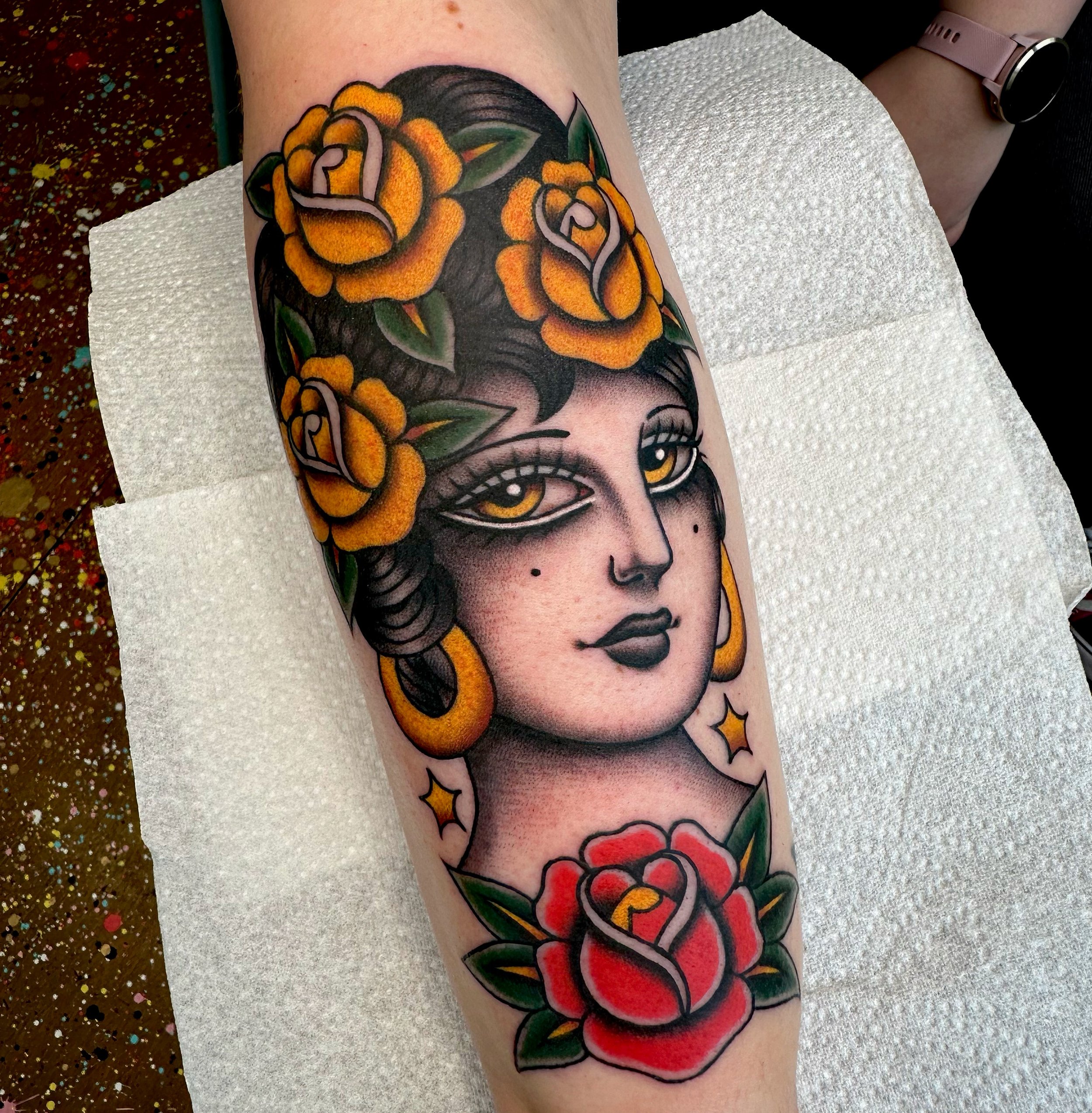 Tattoo of a woman with yellow flowers in her hair, large gold hoop earrings and a red rose.