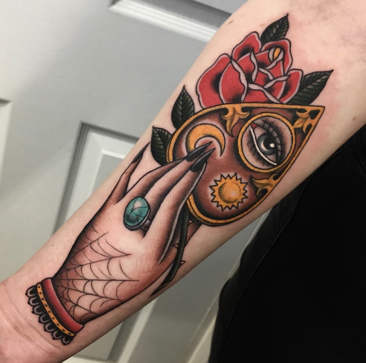 Colorful tattoo of a woman’s hand with a green ring, touching a decorative, ornate face with an eye, sun, and crescent moon. The face has red roses and green leaves above it.