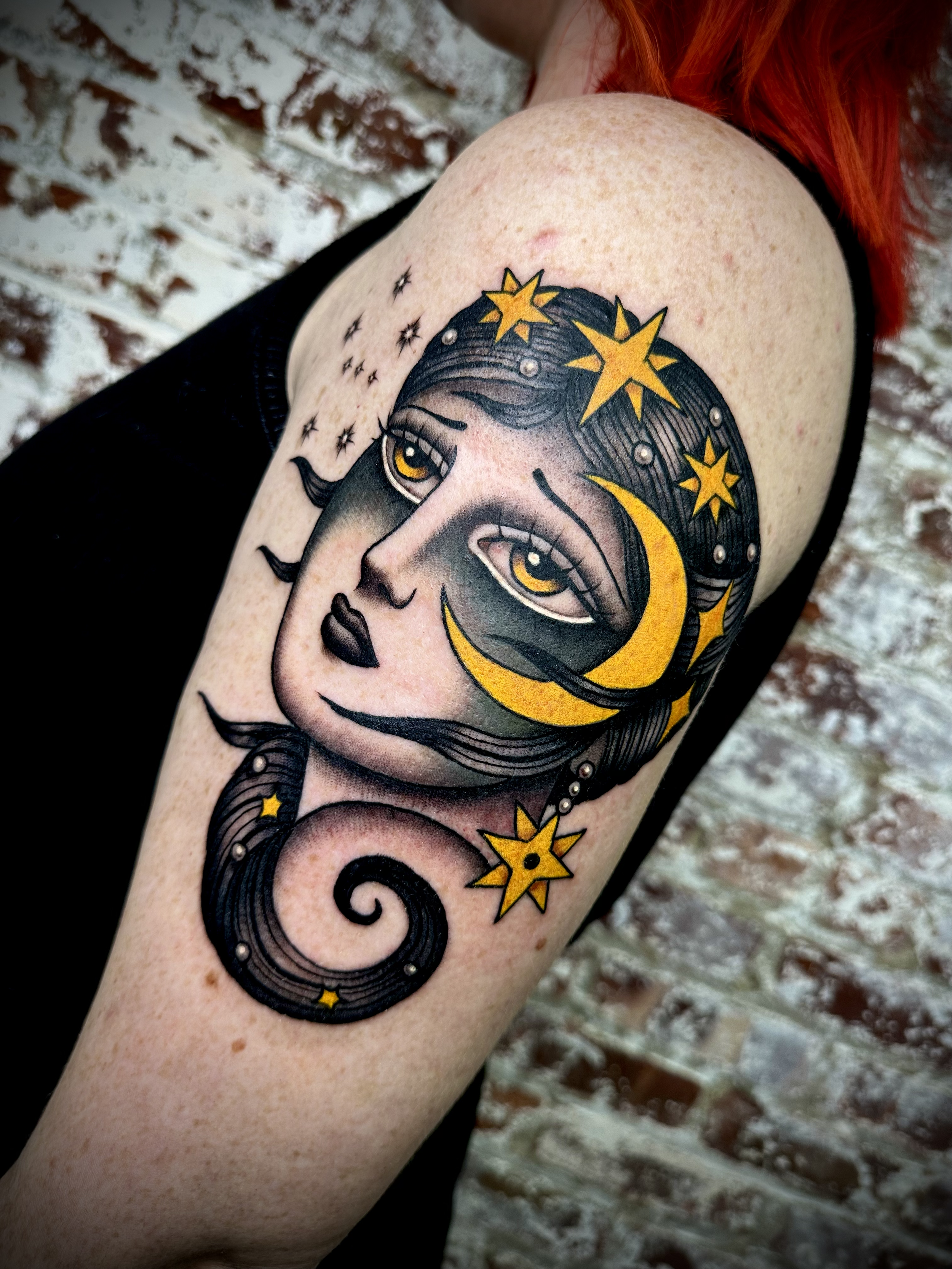 Tattoo of a woman's face with yellow stars and crescent moons on her forehead, surrounded by black swirling lines, on a person's upper arm.