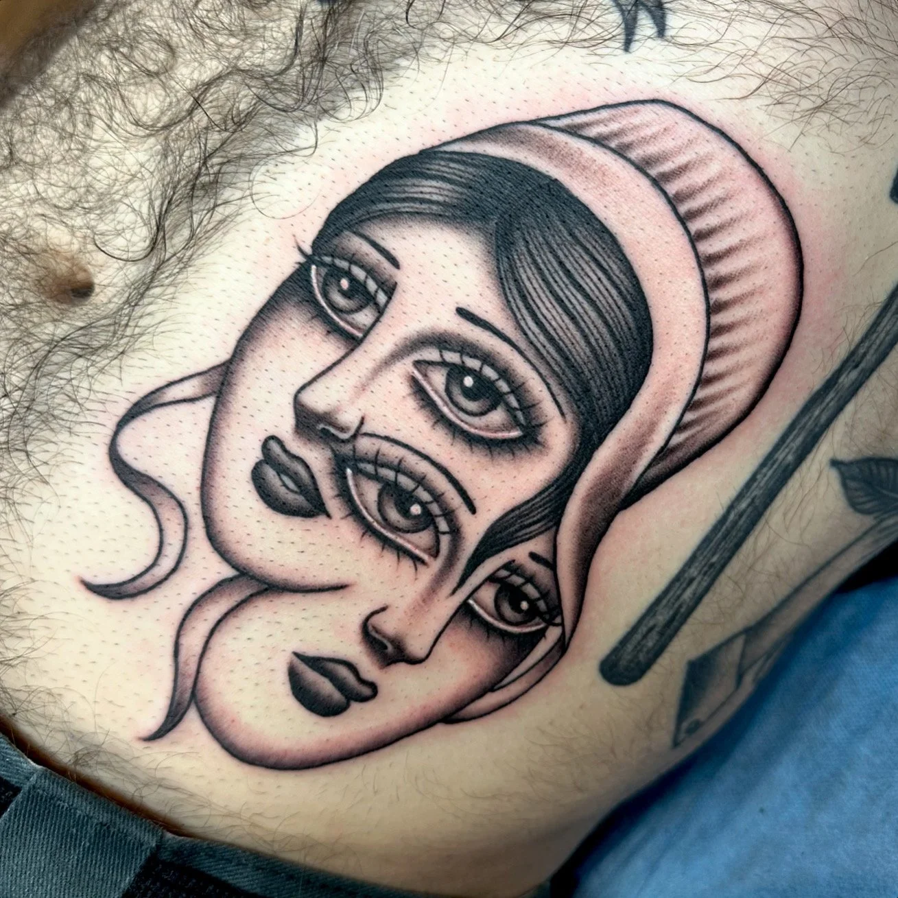 Tattoo of a stylized face with multiple eyes and exaggerated features, including large eyelashes, lips, and a head covering.