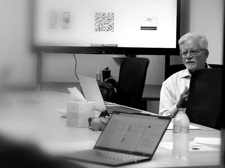 A man with white hair and glasses wearing a white shirt sitting at a conference table in a meeting room, with a large monitor displaying a QR code and other content behind him. The table has laptops, a water bottle, tissues, and other items.