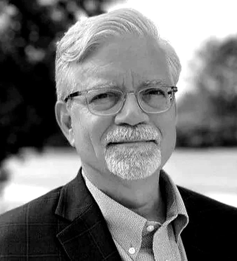 A black and white portrait of an older man with gray hair, glasses, and a beard, wearing a suit jacket and collared shirt outdoors.