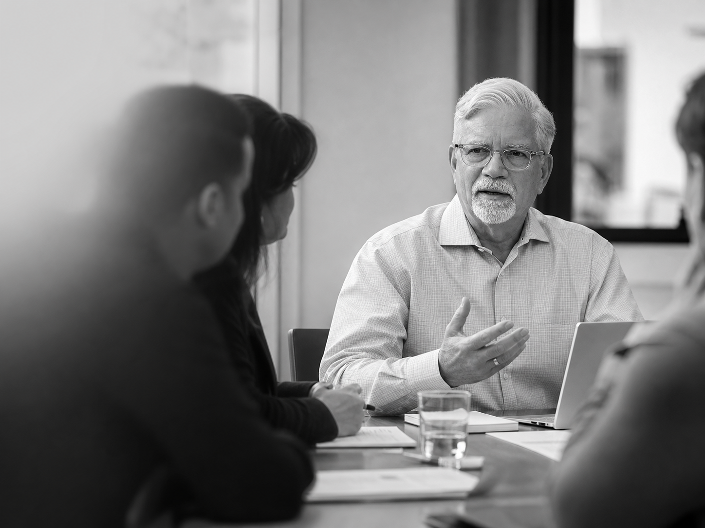 A black and white photo of a man with glasses and gray hair speaking during a meeting with three other people, two of whom are visible, seated around a table with a laptop, papers, and a glass of water.