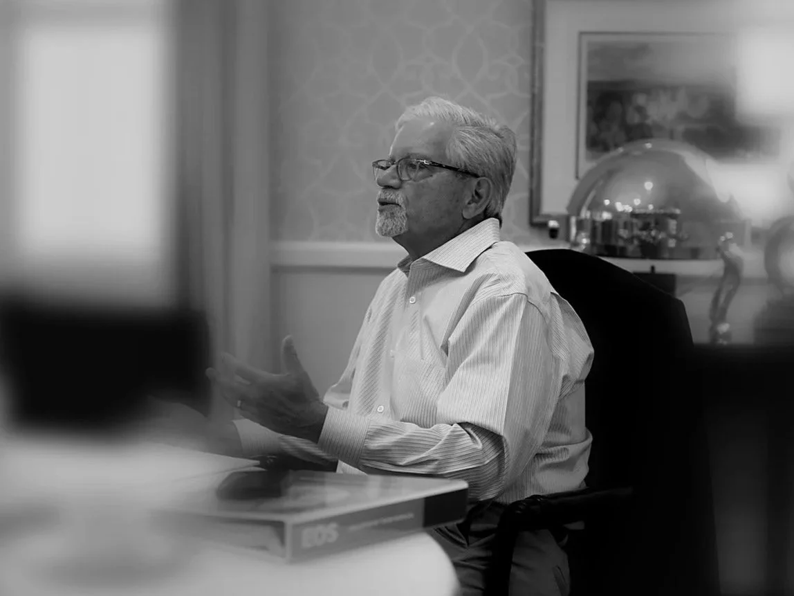 An elderly man with gray hair, a beard, and glasses sitting at a table in a formal setting, gesturing with his right hand while speaking.