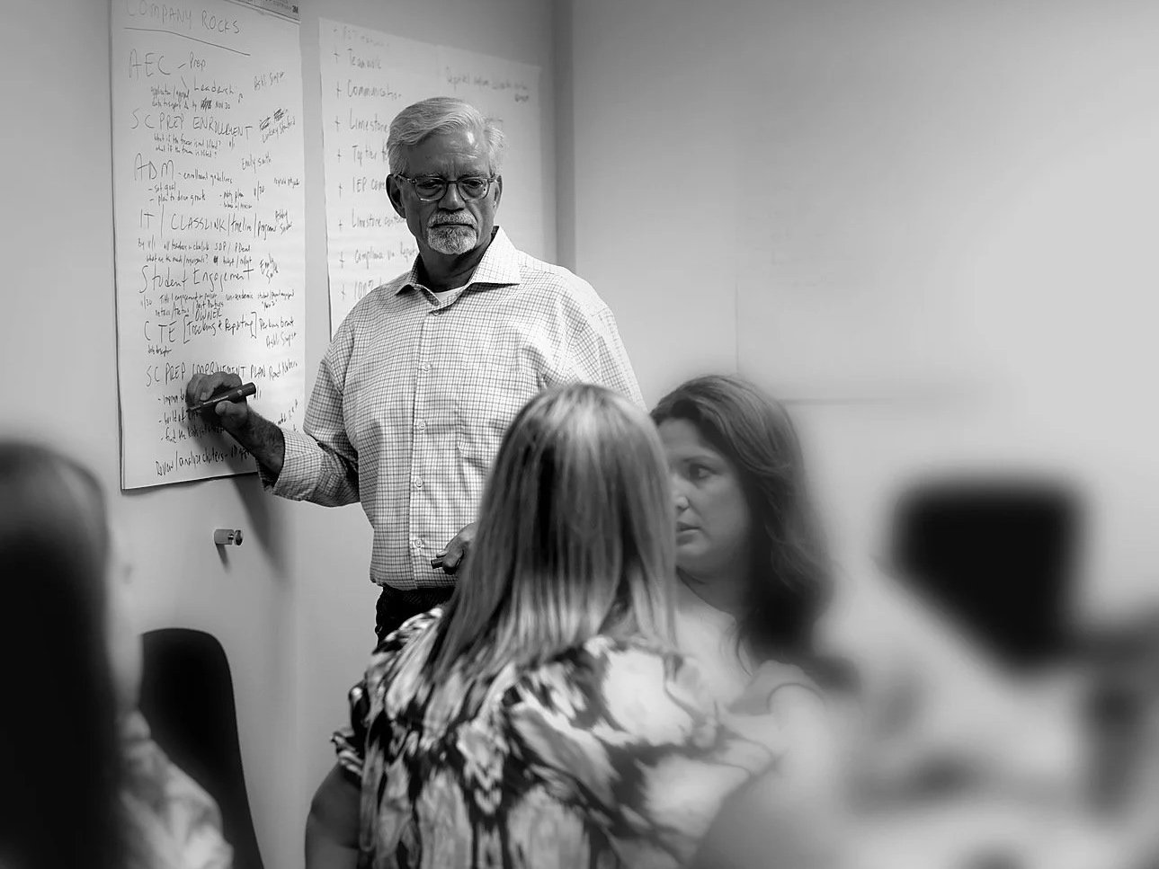 A man with glasses and a checkered shirt stands by a whiteboard, teaching a group of women seated in a room.