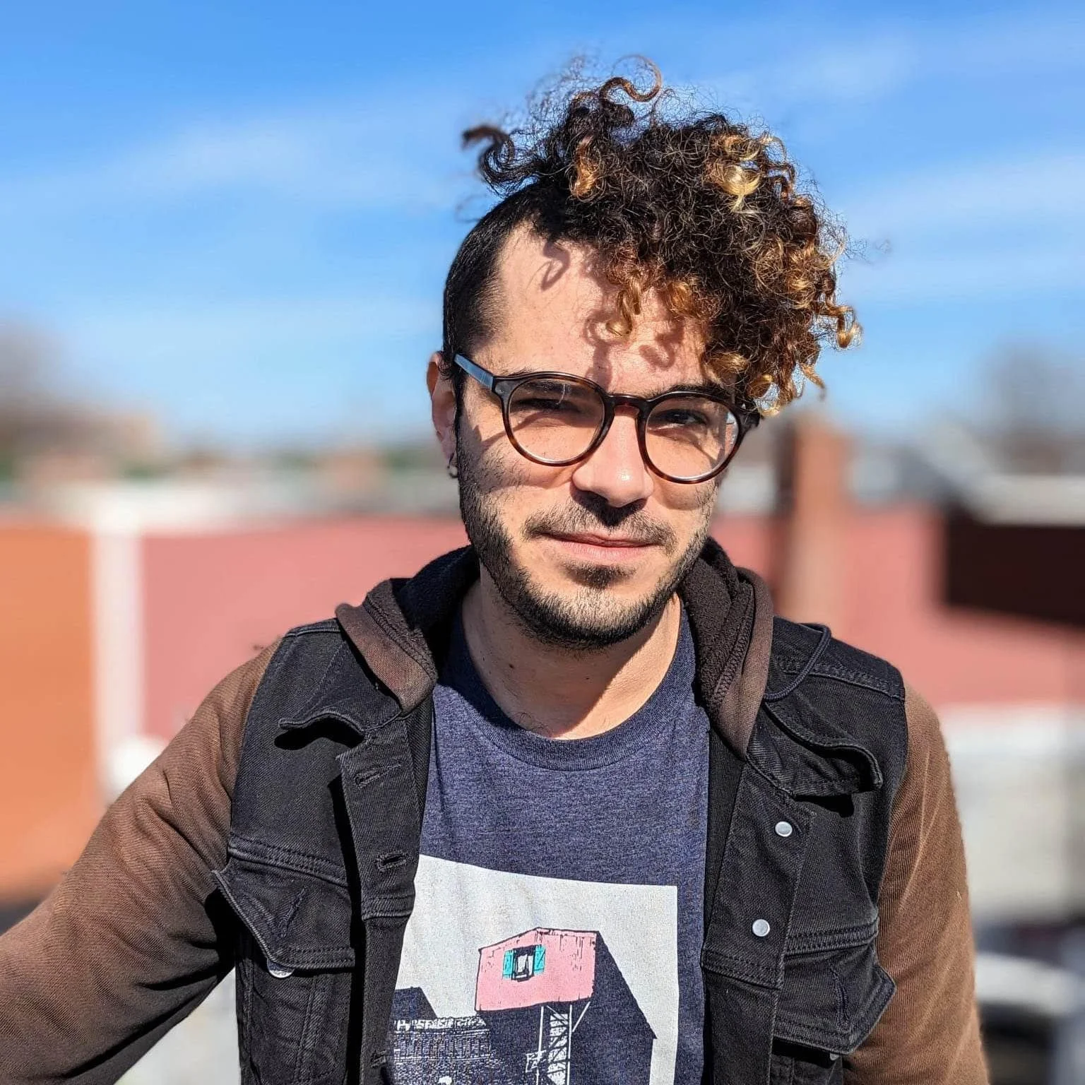 A young man with curly hair and glasses standing outdoors against a blue sky.
