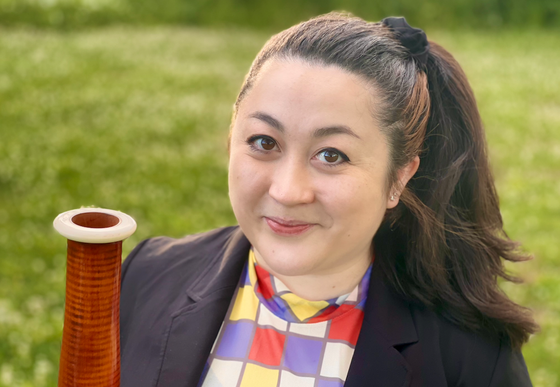 Woman with dark hair tied back, wearing a colorful checkered blouse and dark blazer, holding a wooden didgeridoo outdoors on a grassy field.