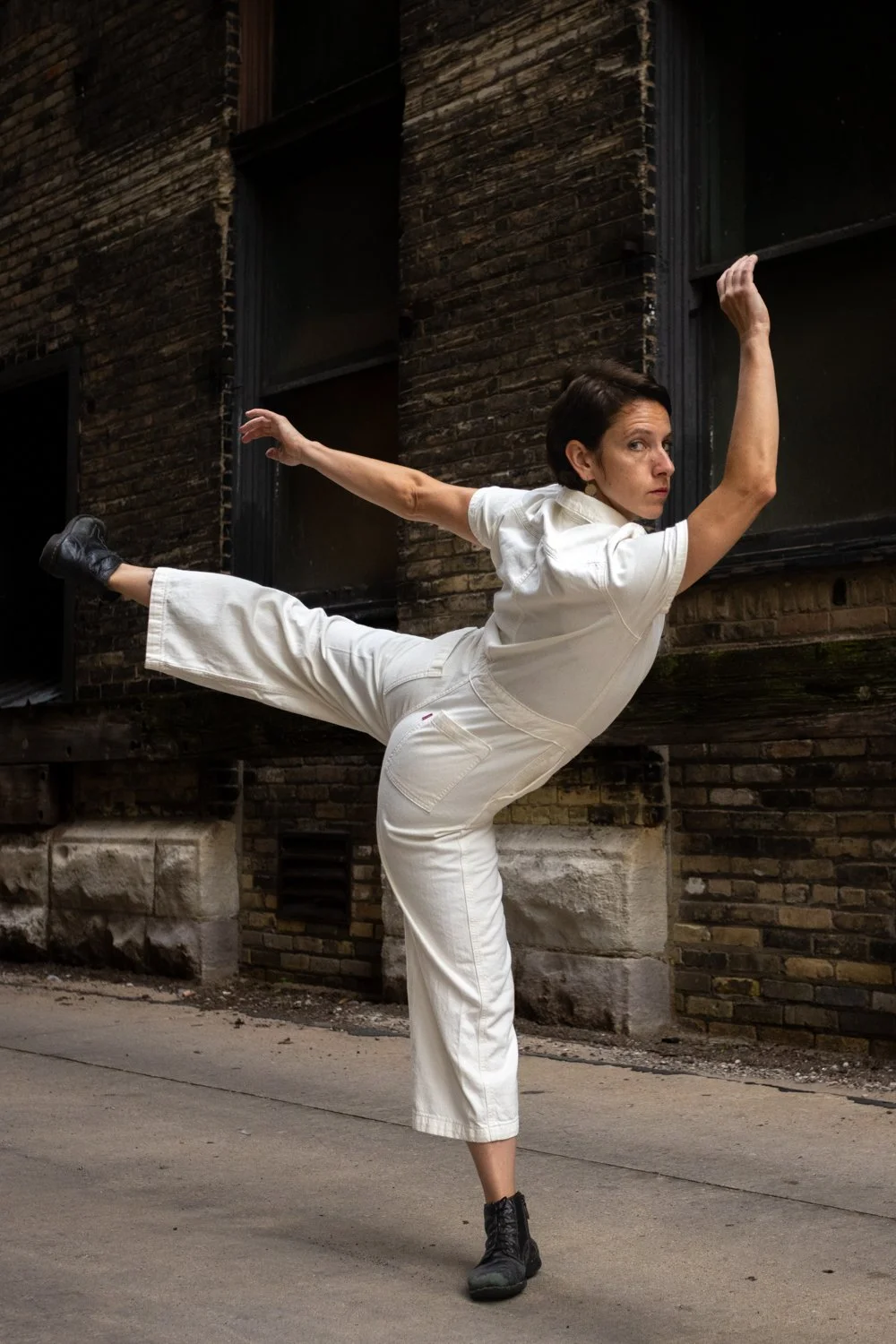 A woman dressed in a white jumpsuit and black boots is performing a high kick against a brick wall in an alleyway.
