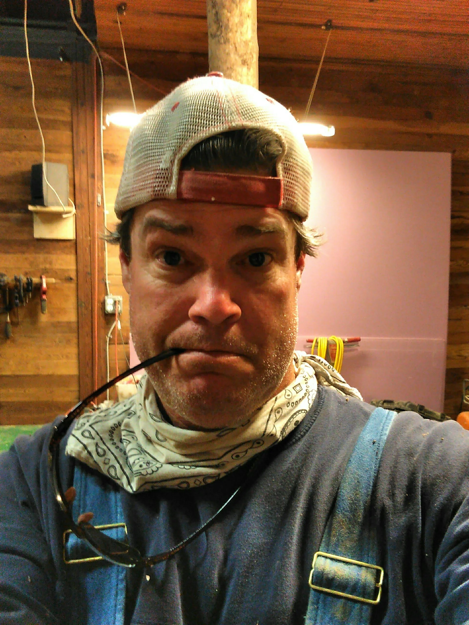 A man taking a selfie in a woodworking workshop, wearing a baseball cap backwards, a bandana around his neck, and safety glasses hanging around his neck.
