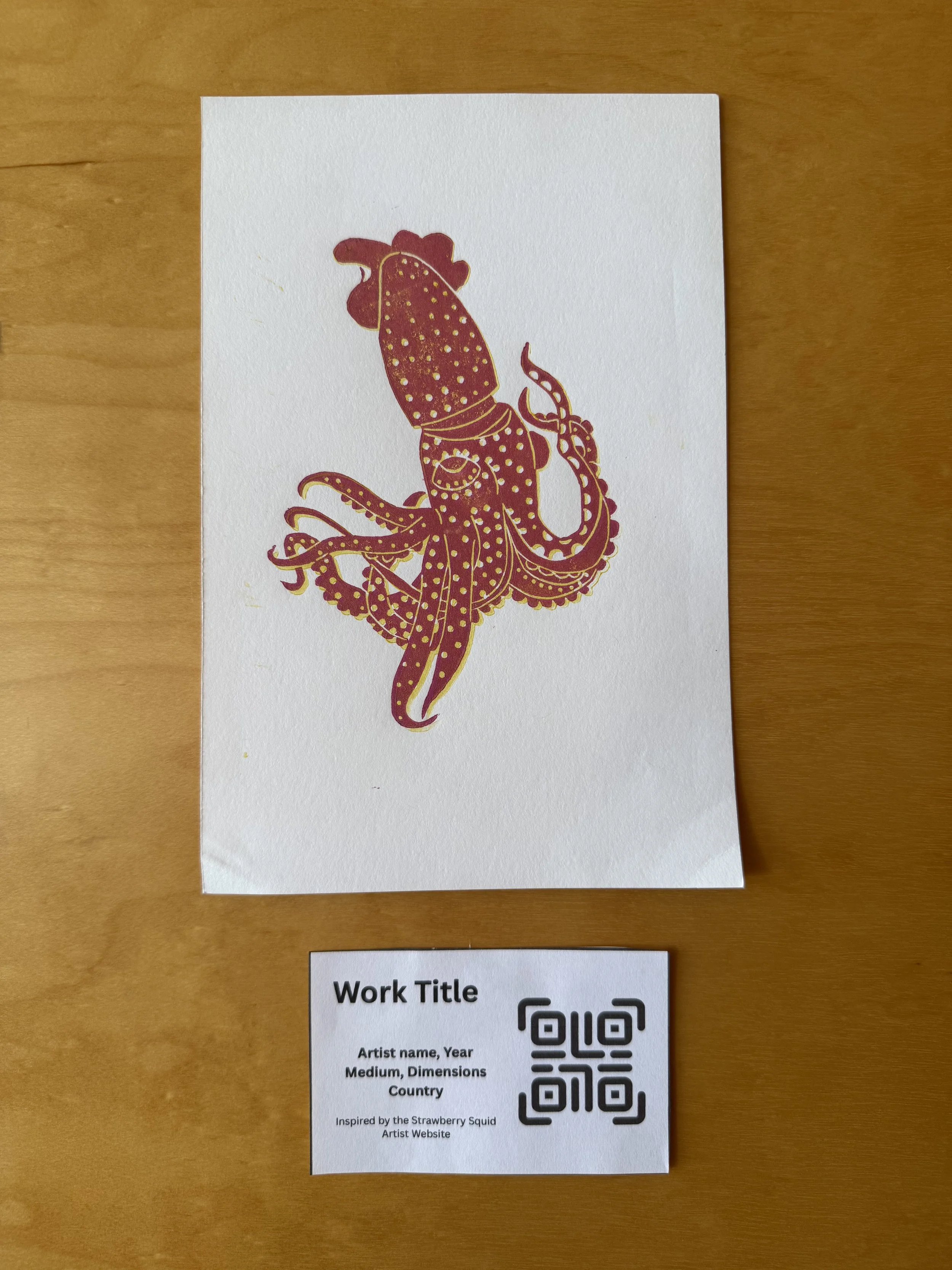 A white sheet of paper with a maroon and gold illustration of an octopus. The paper is on a wooden surface and there is a small label underneath with placeholder text for a work title, artist name, year, medium, dimensions, country, and a QR code.