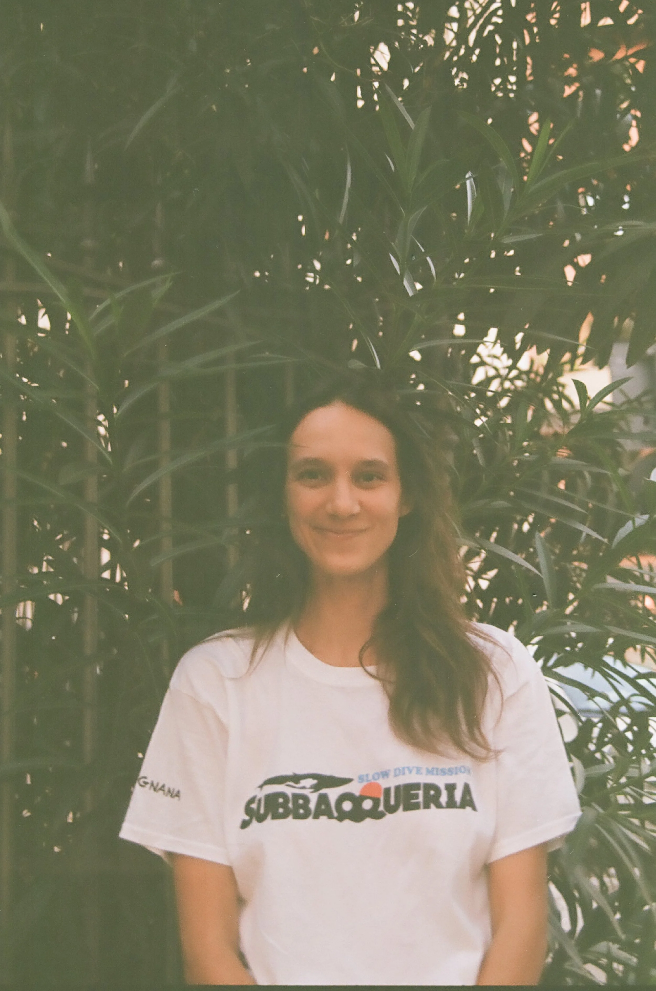 A woman standing in front of large green plants, smiling, wearing a white T-shirt with the logo 'SUBBAQUEERIA' and a small crocodile graphic.