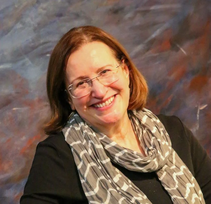 A woman with shoulder-length brown hair, glasses, and a friendly smile, wearing a black top and a patterned scarf, posing against a colorful, abstract background.