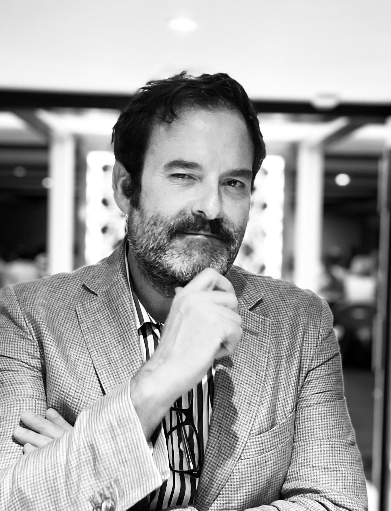 A black-and-white photo of a man with dark hair and a beard, dressed in a checkered blazer and striped shirt, sitting indoors with his hand near his chin and smiling slightly.