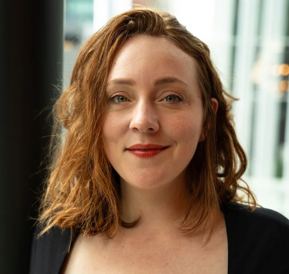A woman with wavy, shoulder-length red hair and fair skin smiling in front of a window.