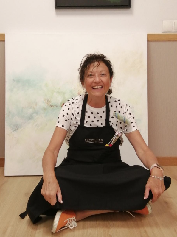 Smiling woman sitting cross-legged on the floor, wearing a black apron and polka dot shirt, with art supplies in her apron pocket, in front of an abstract painting.