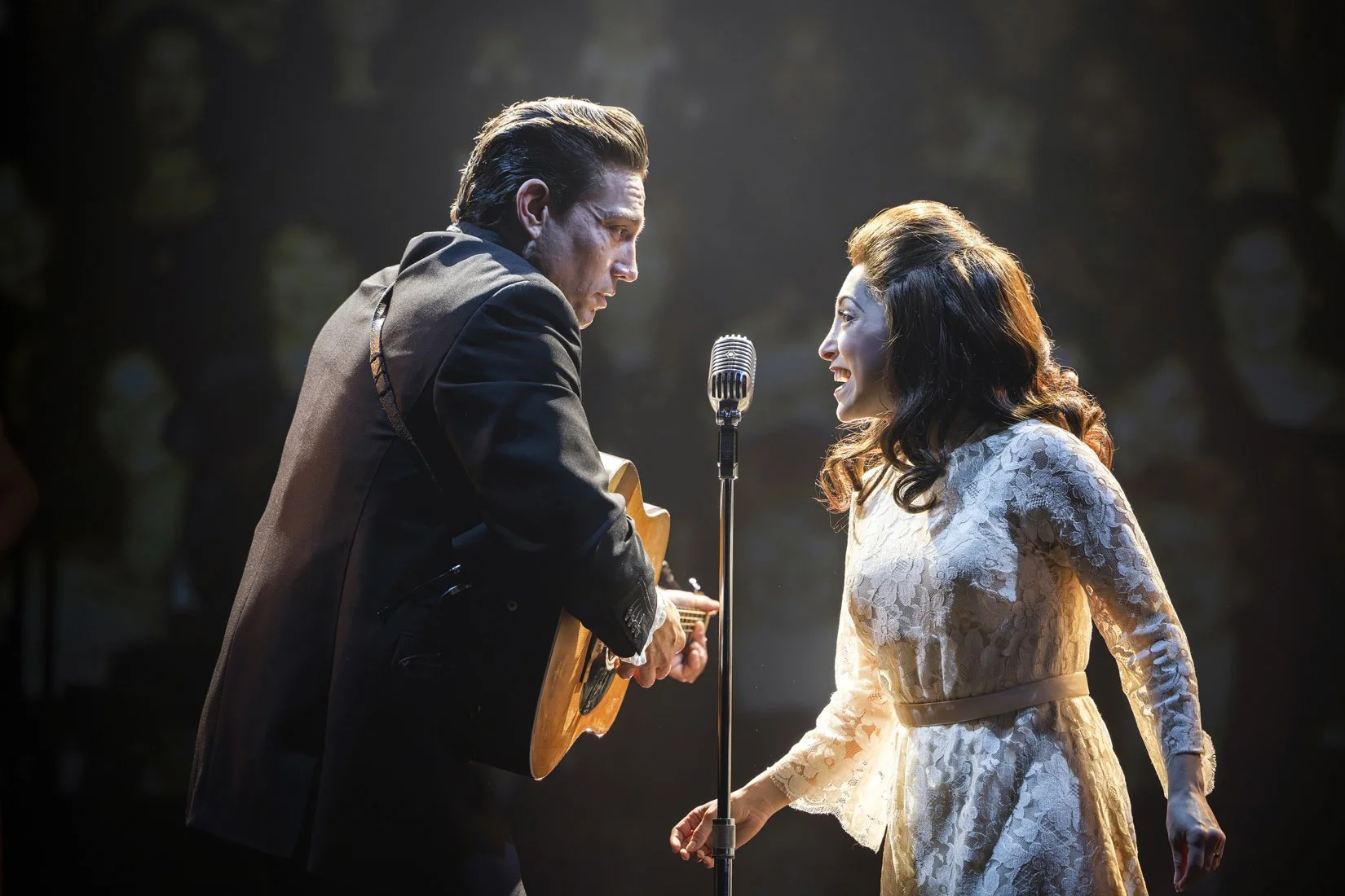 A man and woman perform on stage together, with the man playing guitar and singing into a microphone and the woman smiling at him, wearing a white lace dress.