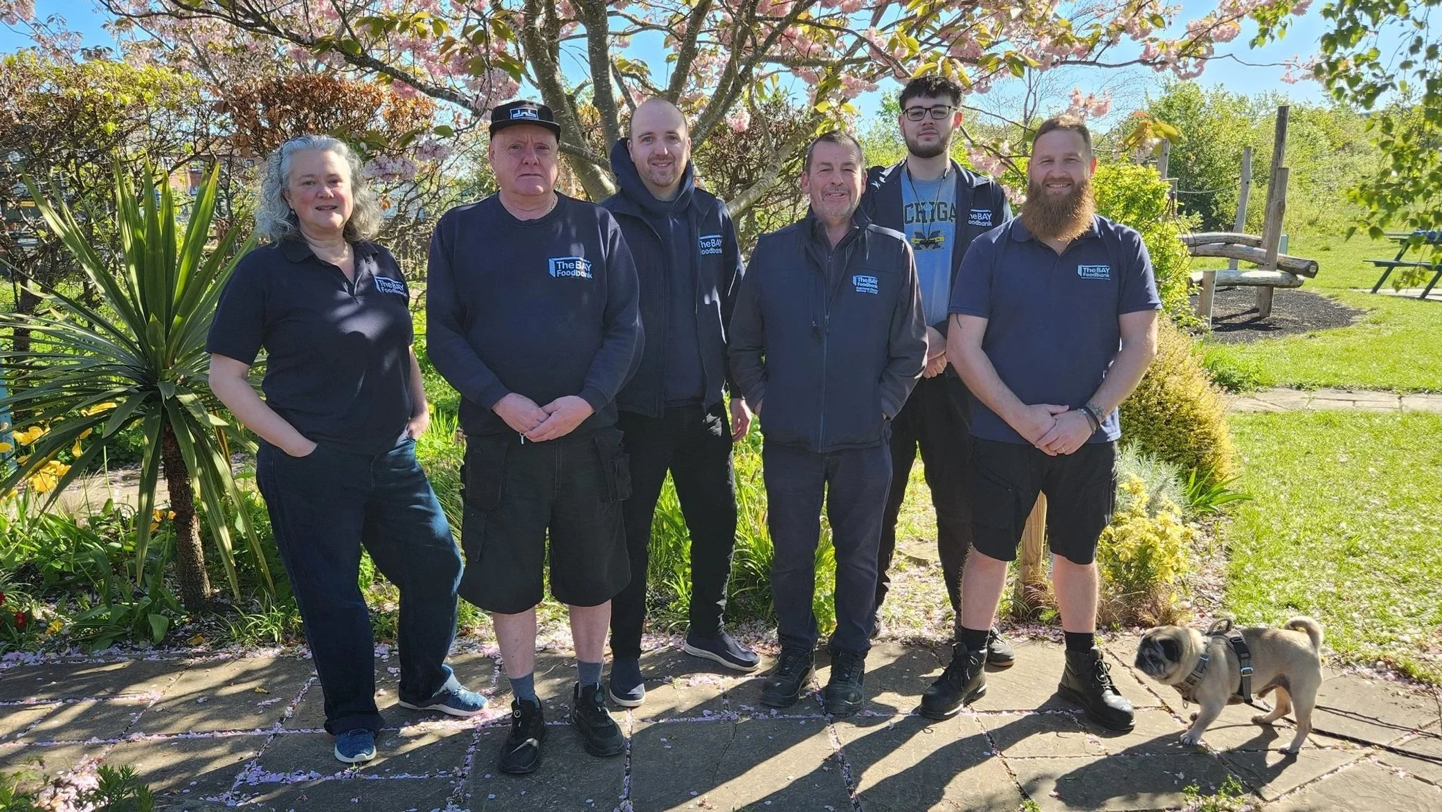 Six people standing outdoors under a blossoming tree with pink flowers, smiling at the camera. A small dog is at their feet on the right. The group wears foodbank uniforom, with 'The Bay Foodbank' logo.