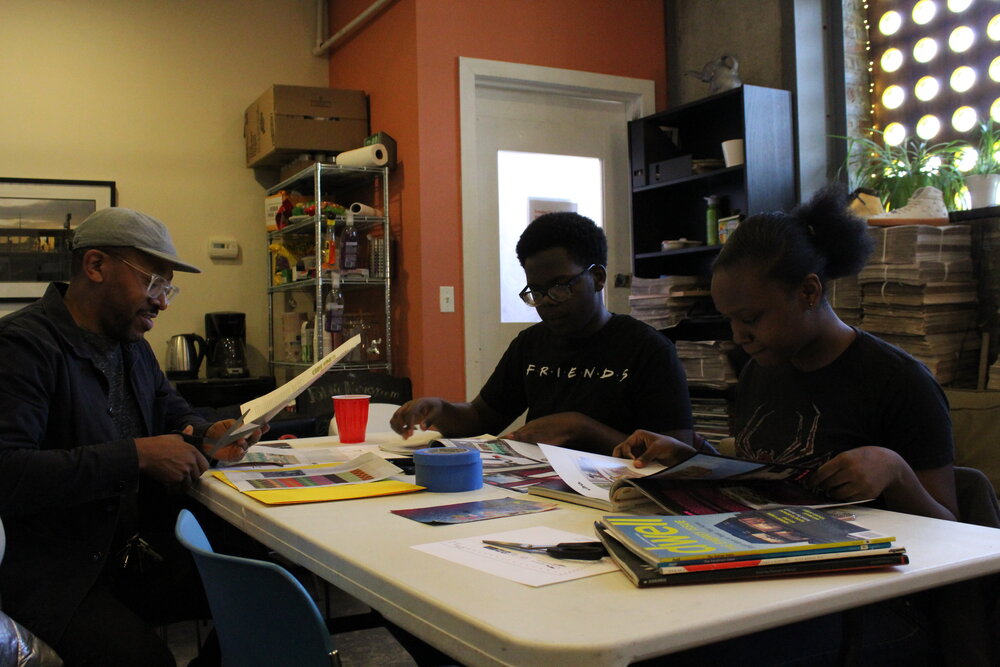  Adult sitting at table with students, all cutting magazines. Credit: Mae Morris 