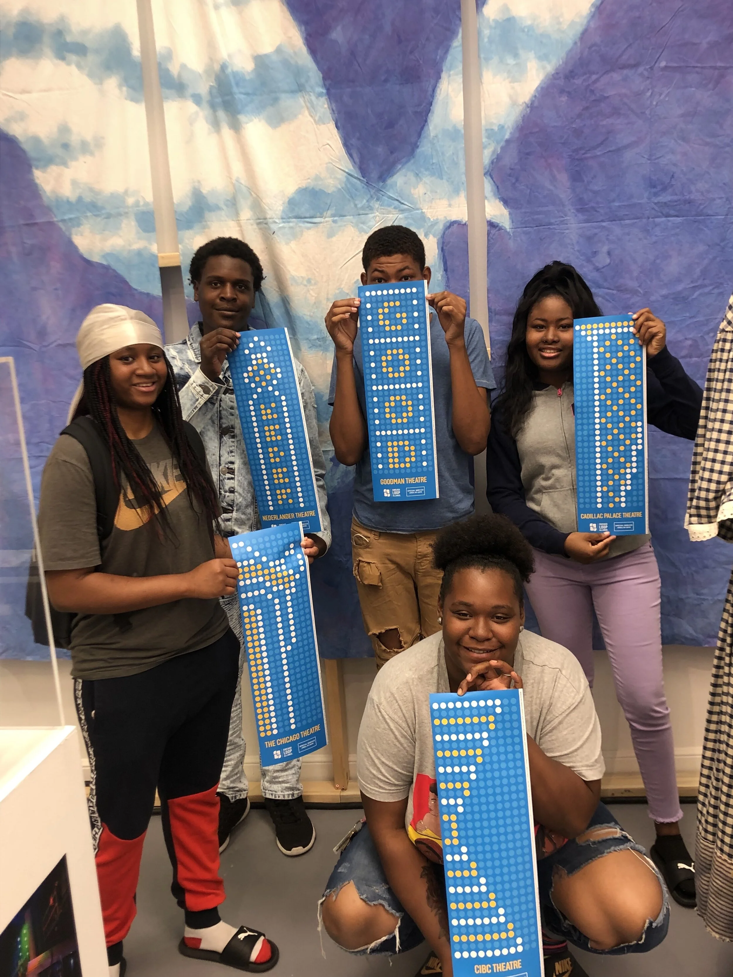 Five young adults holding small replicas of lightpole banners with yellow, white, and blue dots on a blue background that mimic theater marquees.