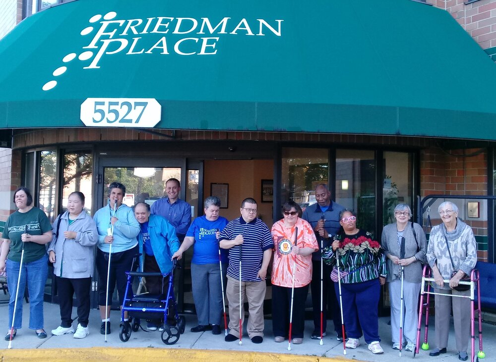  Group of 12 people who are blind or visually impaired standing with canes and walkers under a green awing that reads “Friedman Place” and “5527".” 