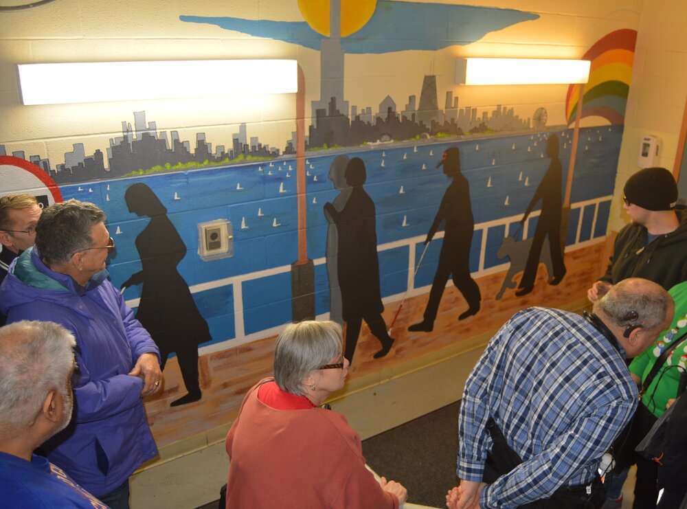  Photograph of people walking along a hallways with a mural of shadows of people who are blind or visually impaired walking on a blue background with the Chicago skyline above their heads. 